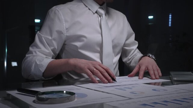 Business professional analyzing financial data with charts and reports on desk, working late in dark office. Audit and overworking concepts
