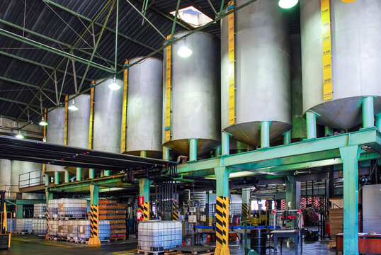 Industrial facility featuring large on a green metal structure, with IBC totes, barrels, and equipment below in a warehouse setting. Focused on chemical or oil processing.