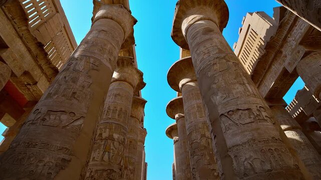 Dramatic low-angle view of enormous carved columns in Karnak Temple Hypostyle Hall, Luxor Egypt. Ancient red painted hieroglyphics on towering sandstone pillars.