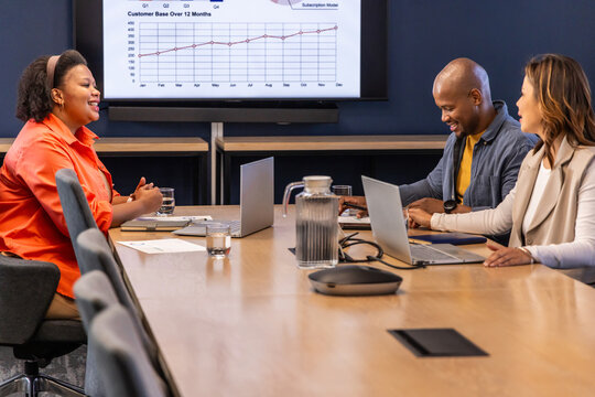 Diverse team meeting around long table in boardroom in business casual with laptops, speaker, chart