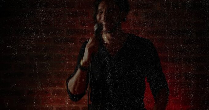 Vertical video: Camera panning man in dark shirt lifting mic cable to mouth by red brick performing