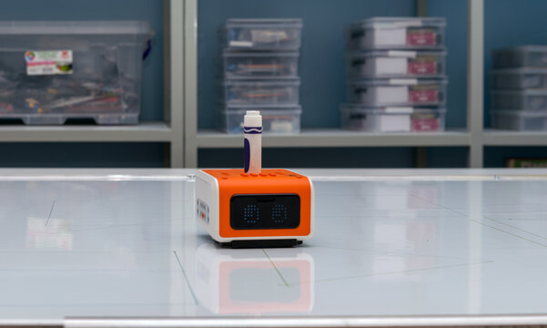 A compact orange and white educational robot toy stands on a glossy reflective white surface in a modern classroom or laboratory environment. The small cube-shaped robot features a black digital LED 