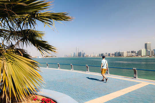 Traveler walking along the coastal promenade in Abu Dhabi, United Arab Emirates. Sunny waterfront scene with palm trees, modern city atmosphere, and views of the Arabian Gulf.