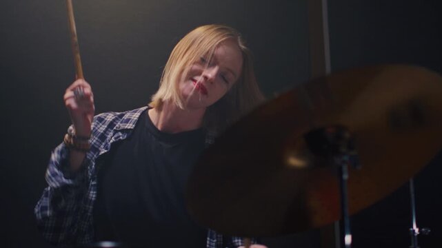 Young girl blonde rock is playing the drums on concert stage. Woman bangs on a drum kit. Face of female musician bangs drumsticks in a snare drum, cymbals in rehearsal studio, repetition. Portrait