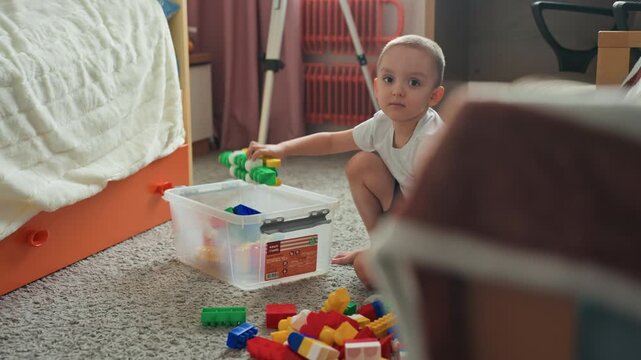 Son building colorful blocks on carpet, small boy kneeling beside clear plastic container, stacking tall tower with focused smile, morning sunlight fills cozy bedroom, creative play supporting fine