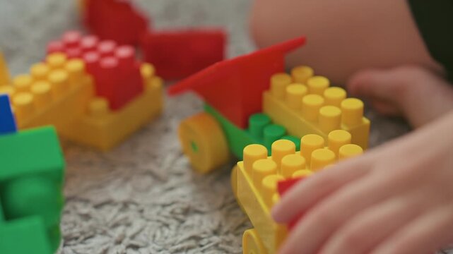 Closeup hands building yellow toy car on carpet, son places wheels and red roof pieces with deliberate grip, tactile engineering play encouraging dexterity and problem solving, warm indoor scene