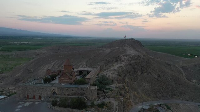 Aerial drone footage flying over the ancient Khor Virap monastery in Armenia. The historic stone complex sits on a hill with the magnificent Mountain in the background.

