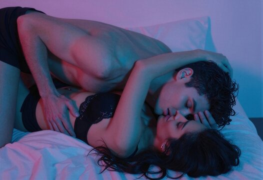 Una pareja se besa apasionadamente sobre una cama blanca iluminada con luces de ne&oacute;n.