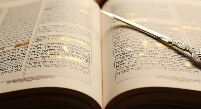 Open jewish holy book with golden hebrew text and a decorative silver yad or pointer, used for reading from the torah during religious services.