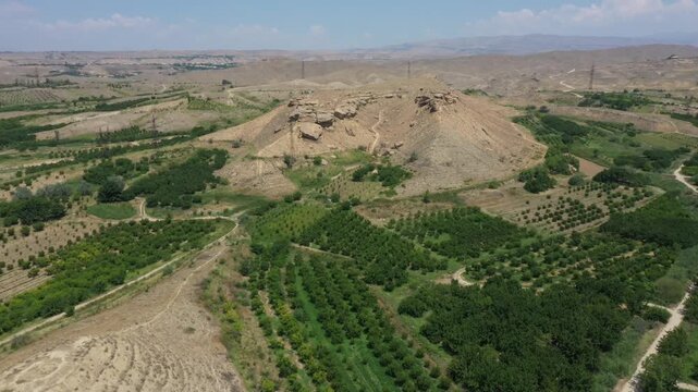 Aerial drone footage of a mysterious ancient stepped earthen pyramid, possibly a burial mound or kurgan, beautifully situated amidst lush agricultural fields and orchards in a scenic valley.

