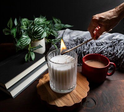 Hand lighting a candle in a cozy home environment with tea, books, and a soft blanket. This lifestyle moment captures a calming evening ritual, representing mindfulness, relaxation, and intentional li