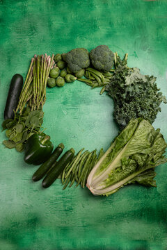 Assorted fresh green vegetables arranged in a circular composition on a textured green background. Includes broccoli, asparagus, kale, zucchini, green beans, spinach, and lettuce, representing healthy