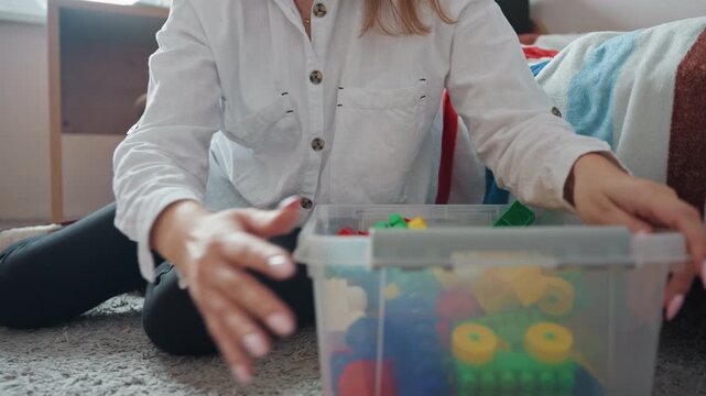 White mom tidying sons colorful blocks, kneeling on carpet beside plastic storage bin, hands reaching into box to sort large interlocking pieces, casual white shirt and black leggings, soft natural