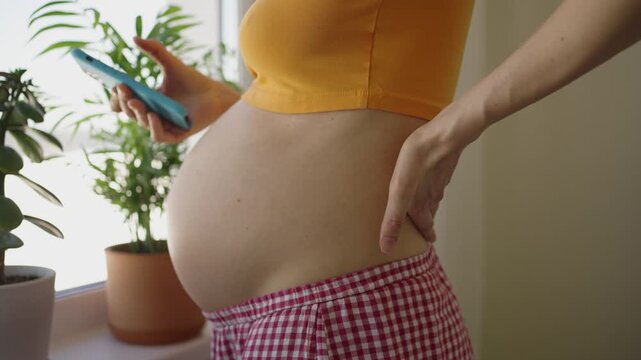 Expecting mother standing indoors with a large bare belly, holding her lower back in discomfort while using a smartphone, illustrating the physical strain and pain during late-stage pregnancy