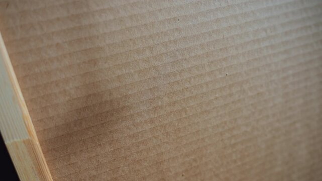 Closeup cardboard surface with dent showing corrugation and texture. Soft side lighting reveals subtle crease near box edge, fine fiber detail, light scuffing and faint stain. Neutral brown tones