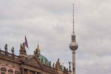 Obraz premium Berlin TV Tower stands tall against cloudy sky. Historic building with ornate roof and flag flies beside tower. No people visible. Urban skyline image ideal for travel, architecture