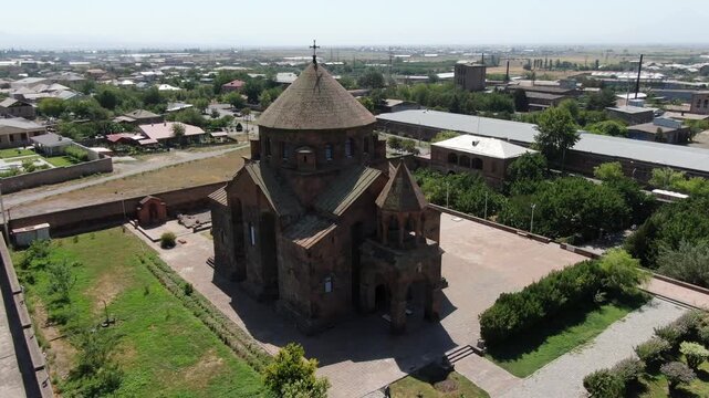 Aerial drone footage circling a magnificent ancient Armenian church made of stone with a distinctive conical dome, situated in a town surrounded by trees on a sunny day.

