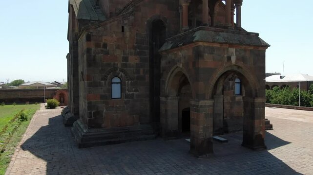 Aerial drone footage circling a magnificent ancient Armenian church made of stone with a distinctive conical dome, situated in a town surrounded by trees on a sunny day.


