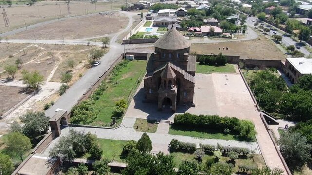 Aerial drone footage circling a magnificent ancient Armenian church made of stone with a distinctive conical dome, situated in a town surrounded by trees on a sunny day.

