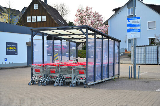 ISERLOHN, GERMANY - MARCH 27, 2026: Rows of red shopping carts in a covered outdoor storage bay at a Penny discount supermarket parking lot, surrounded by suburban residential houses.