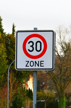 Speed limit 30 zone sign on a pole with trees and houses in the background