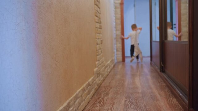 White son running down hallway past bricktrimmed wall on wooden floor cat watches from left, child in white shirt and dark pants, slippers visible, playful rush toward doorway while moms voice heard