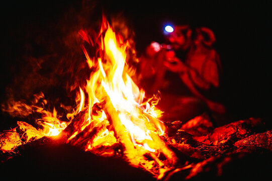 A man wearing a headlamp eats food near a blazing campfire