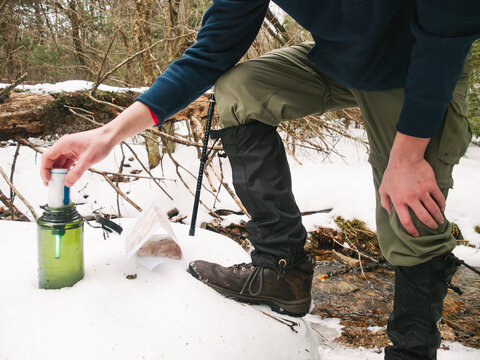 A young man filters water from a stream into a water bottle using a ultraviolet-light filter while backpacking in winter.