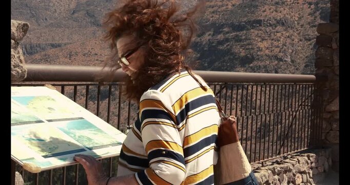 Senior woman reading an information sign at a tourist destination while the wind moves her hair. Concept of active aging, independence and enjoying travel during retirement.