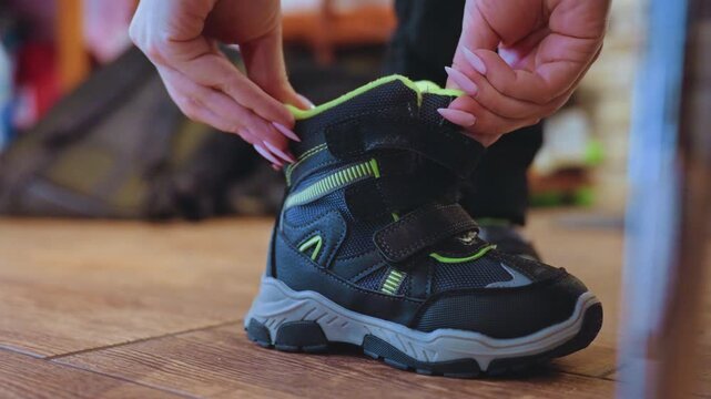 Mom tying sons winter boot, gentle hands fastening velcro strap on black green toddler boot on carpet, morning routine for school, cozy home interior, closeup caring parent preparing child, practical