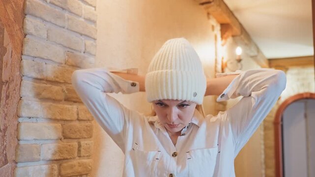 Woman adjusting knit beanie in hallway, tugging plush coat over shoulders, focused expression, warm brick wall lighting, cozy indoor entryway, slow deliberate movements, neutral shirt, soft textures,