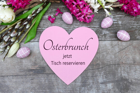 Rosa Herz mit dem Text 'Osterbrunch jetzt Tisch reservieren' auf Holztisch umgeben von Fr&uuml;hlingsblumen und dekorativen Ostereiern.
