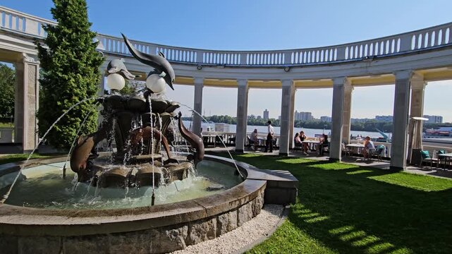 MOSCOW - JUL 26 , 2025: Fountain with a statue of a dolphin in the middle. The fountain is surrounded by a grassy area and a walkway. There are several people in the area, tables and others standing