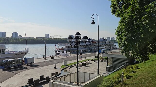 MOSCOW - JUL 26 , 2025: City street with a river running through it. The street is lined with trees and has a few benches. A boat is docked in the river Stalin era riverside station
