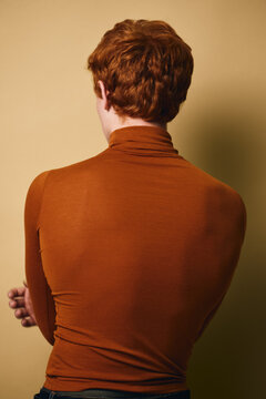 Person with red hair facing away in brown turtleneck sweater against beige background. Casual style with folded arms and short hairstyle showing back view indoors.