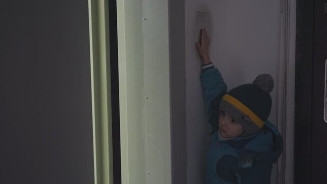 Son at doorway reaching for intercom. Small white boy in winter coat and striped beanie presses wall intercom while waiting in dim hallway graycoated white mom approaches from behind, gentle