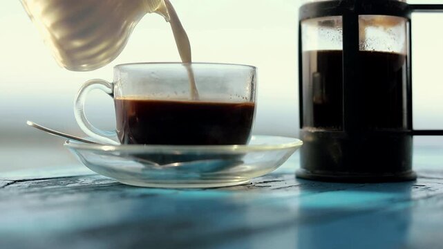 Pouring cream into Balinese coffee and stirring with a spoon, next to a coffee pot on a blue table.