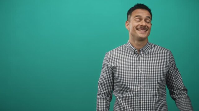 Man with moustache in checkered shirt smiles and gives thumbs up in studio with teal backdrop; cheerful confidence.