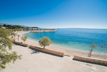 Obraz premium Wide-angle view of Znjan beach in Split, Croatia. Scenic coastline with stone benches, green trees, and crystal clear blue Adriatic sea under a bright sunny sky.