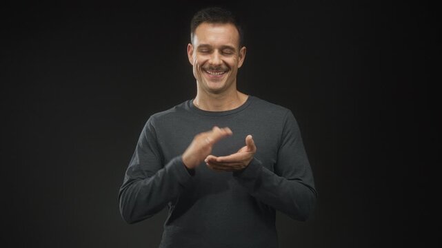 Man with moustache claps hands in studio while smiling at camera, wearing dark gray sweater and looking pleased; celebration joy.
