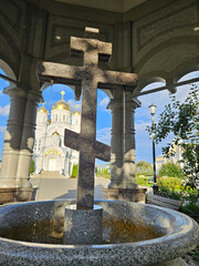 Fototapeta premium Large stone cross is in the middle of a fountain. The fountain is surrounded by stone pillars and a church is in the background