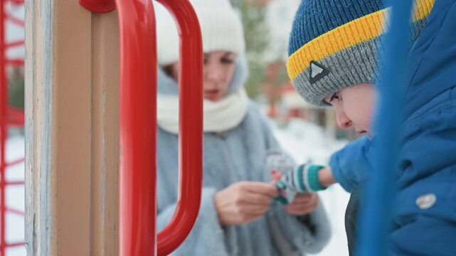 Mom helps son on playground winter, fastening glove on son in blue jacket and knit hat with yellow stripe, red metal rail in foreground, mother adjusting cuff with focused gentle expression, chilly