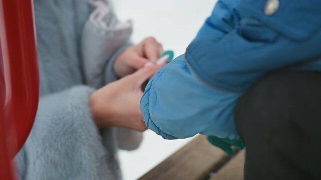 Closeup hands putting sock on son, winter bench scene mom gently pulls striped knitted sock onto childs foot, blue coat and wooden bench, snow in background, warm caring interaction and intimate