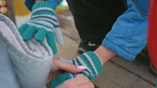 Mom helping son put on mittens, intimate closeup of hands fastening striped knit gloves on wooden playground deck, mother gently guiding childs fingers, winter jacket sleeve, soft touch, caring family