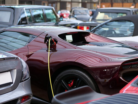  luxury maroon Ferrari 296 GTB plug-in hybrid
charging at a showroom. A bright yellow cable is connected to the side
charging port. Surrounded by a collection of classic and modern luxury
vehicles 