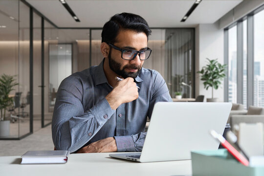 Young serious indian professional business man, focused ethnic male student wearing glasses working on laptop, remote studying using computer looking at screen watching seminar webinar at home office.
