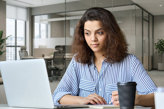 Young indian female student working on laptop in modern office coworking space classroom. Hispanic business woman using computer watching virtual online training course, remote learning concept.