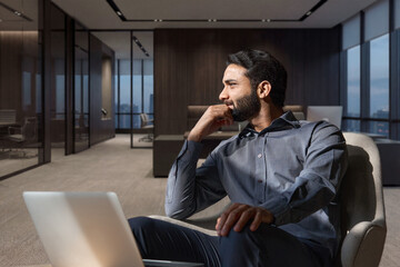 Young thoughtful smart indian professional business man executive looking away relaxing sitting on...