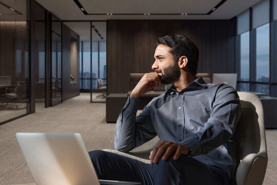 Young thoughtful smart indian professional business man executive looking away relaxing sitting on chair in modern office lobby with laptop, thinking of new ideas, dreaming of success, planning.