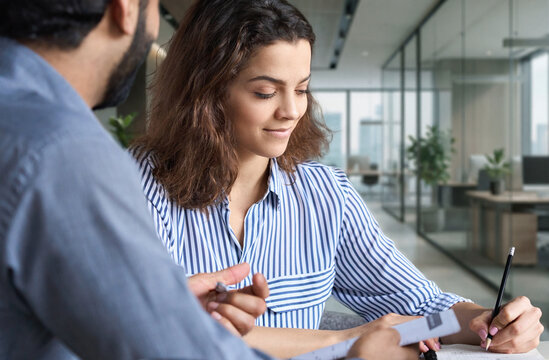 Two sales agents colleagues working together with papers in office. Indian male mentor and latin female young professional talking in creative office space.
